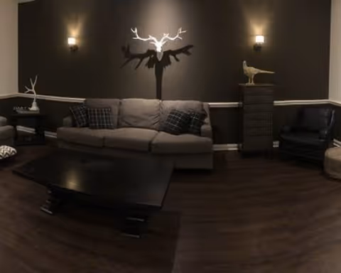 A cozy living room with a gray sofa adorned with plaid pillows, a dark wooden coffee table in front, a black armchair to the right, and decorative items including a white antler wall light casting a shadow and a bird sculpture on a tall chest of drawers against a dark wall.
