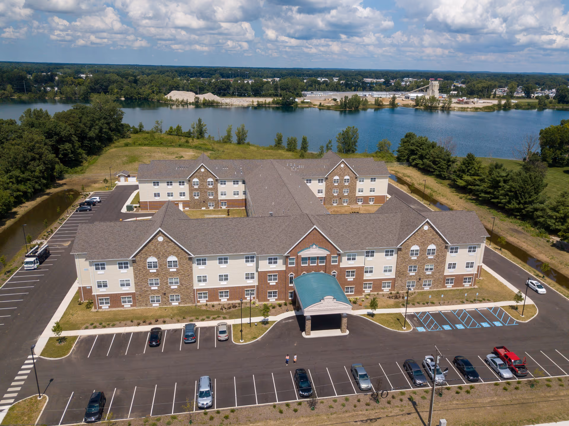 Aerial view of a three-story senior living building with a covered entrance, parking lot, and a river behind it.