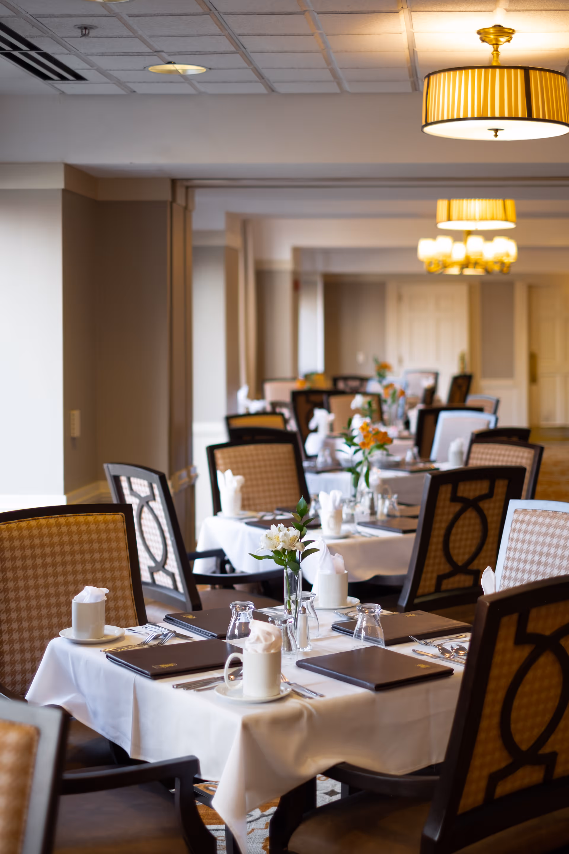 A neatly arranged dining room with tables covered in white tablecloths, set with menus, cups with napkins, glasses, and silverware. Each table has a small vase with flowers. The room has patterned chairs and warm lighting from ceiling lamps.