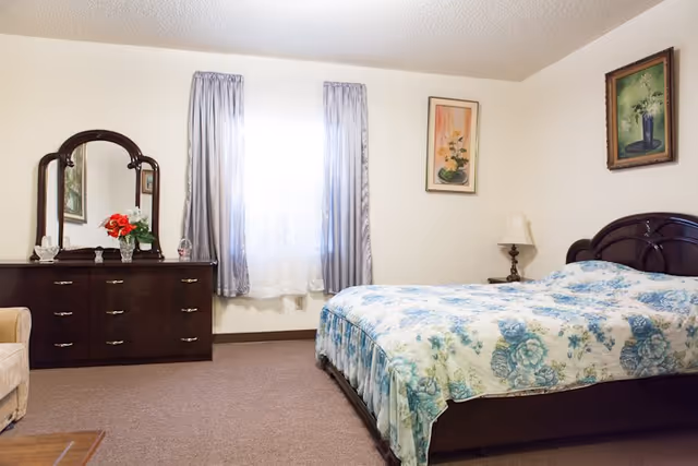 A neatly arranged bedroom with a floral-covered double bed, dark wood dresser and mirror, window with curtains, and framed paintings on the walls.