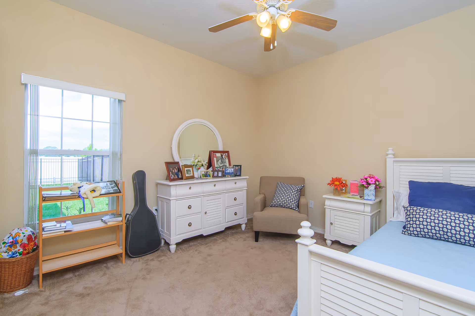 A cozy bedroom with beige walls and carpet. The room features a white wooden bed with blue bedding and patterned pillows, a matching white nightstand with flowers and a book, a beige armchair with a patterned cushion, and a white dresser with a round mirror and framed photos. A guitar case leans against the dresser, and a small wooden shelf stands by a window with vertical blinds, showing a green lawn outside.