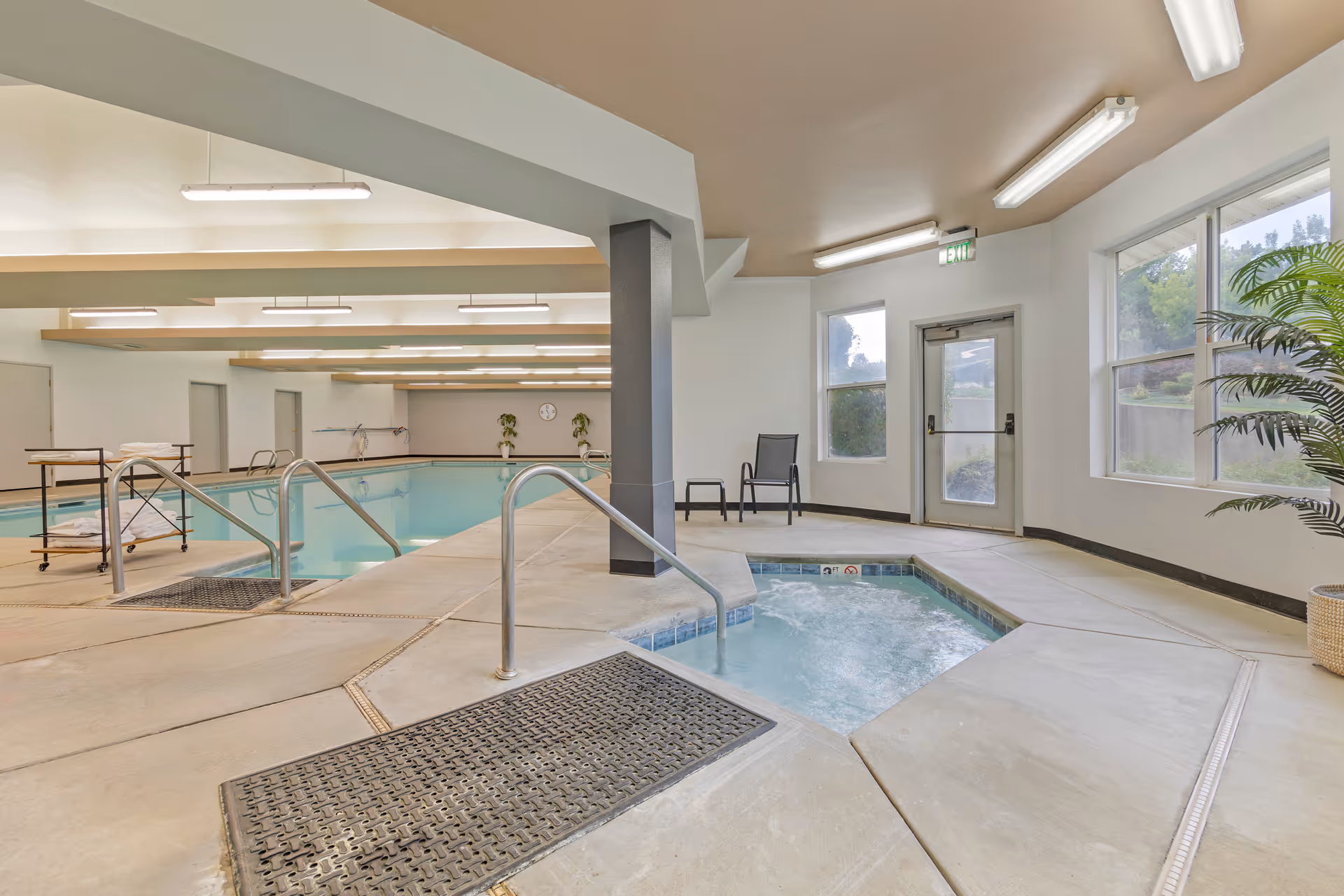 Indoor swimming pool area with a hot tub in the foreground, metal handrails, a chair and small table near windows, and a door leading outside. The pool area is well-lit with ceiling lights and has a clean, spacious concrete floor.