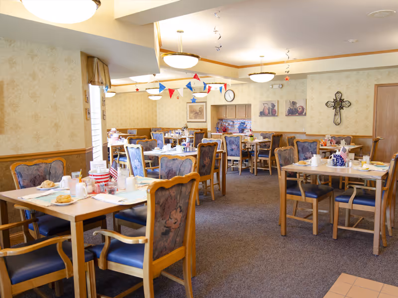 A dining room in a senior living facility with multiple wooden tables and chairs arranged neatly. The tables are set with plates, cups, and utensils, and some have small American flags and patriotic decorations. The room has beige wallpaper with a subtle pattern, framed artwork on the walls, and ceiling lights. Red, white, and blue bunting and star decorations hang from the ceiling.