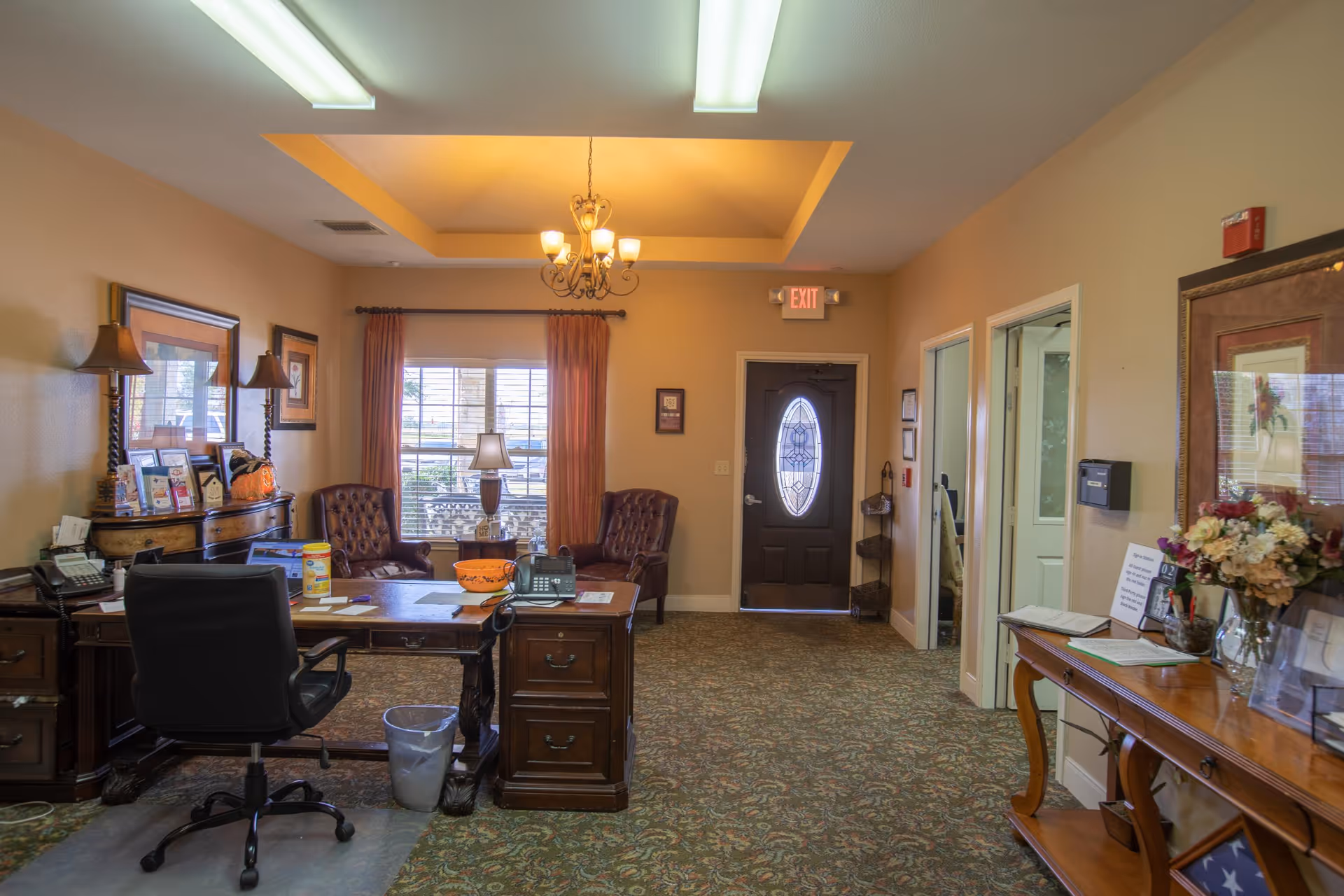 An office area in a senior living facility with a wooden desk, office chair, two leather armchairs near a window with curtains, a chandelier on the ceiling, and a door with an oval glass window. The room has carpeted flooring and beige walls with framed pictures and a table with flowers and documents.