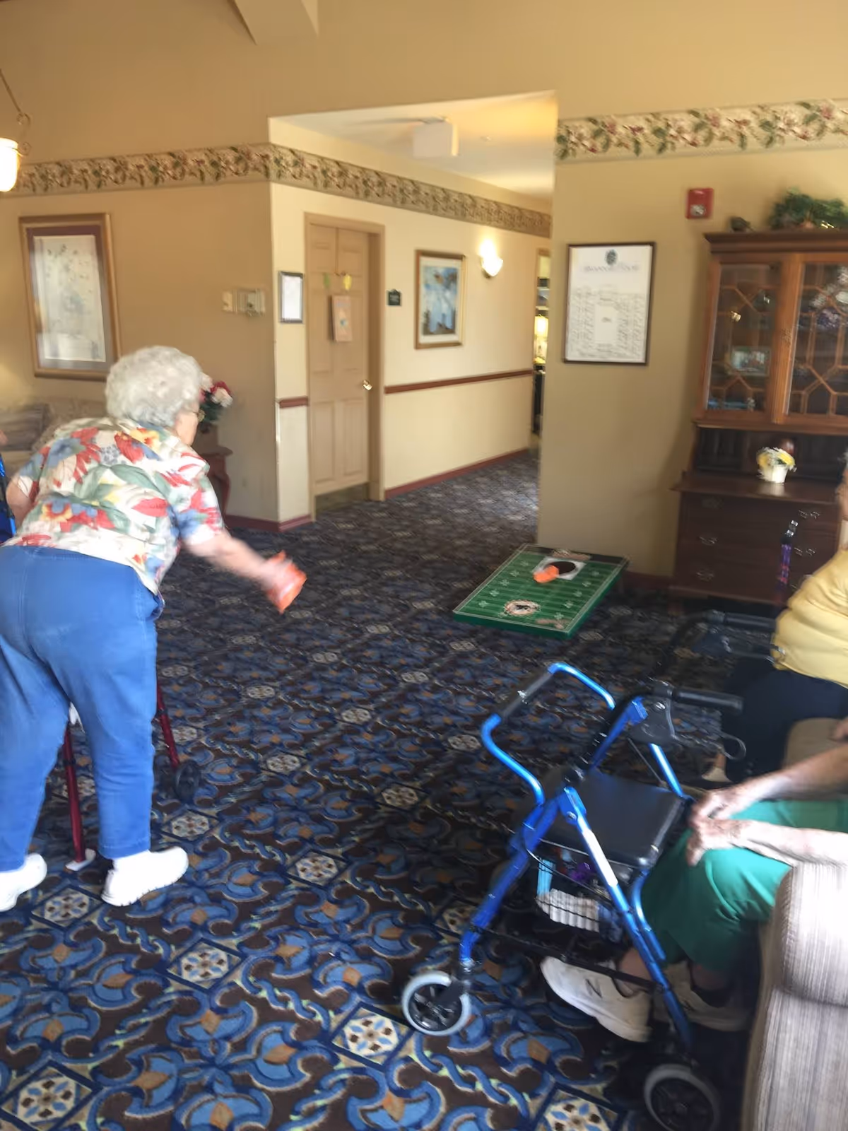 An elderly woman playing a bean bag toss game indoors on a patterned carpet. Another elderly person is seated nearby with a blue walker. The room has beige walls with floral wallpaper border and framed pictures, and a wooden cabinet with glass doors.