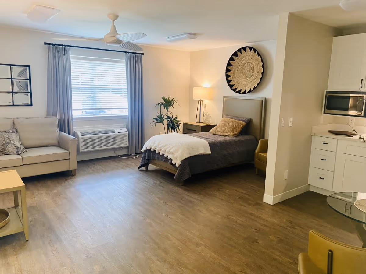 A bright and spacious room featuring a single bed with a gray bedspread and a white throw blanket, a beige sofa with patterned pillows, a window with gray curtains and blinds, a ceiling fan, a small nightstand with a lamp, a decorative wall hanging above the bed, a potted plant, and a kitchenette area with white cabinets and a microwave.