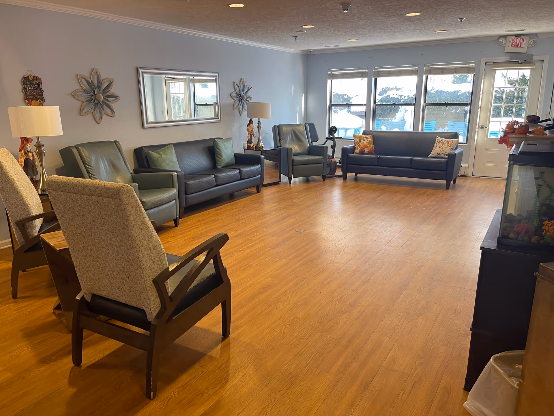 A spacious senior living common area with wooden flooring, two green leather sofas, two green armchairs, and two beige cushioned chairs arranged around the room. The walls are decorated with a large mirror and flower-shaped wall art. There are two table lamps on side tables, a fish tank on the right, and large windows letting in natural light. A door with a 'Not an Exit' sign is visible at the back.