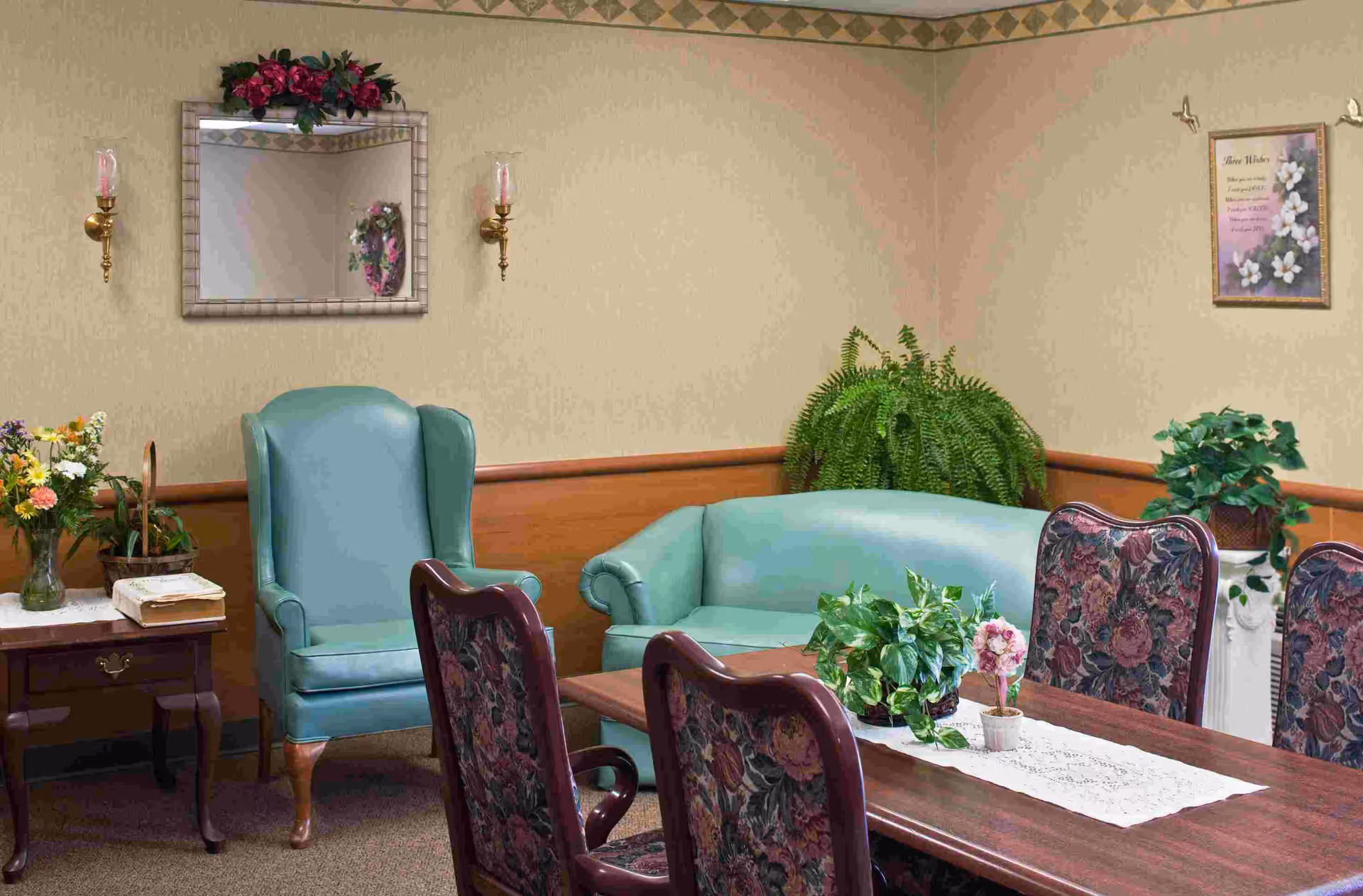 A cozy sitting area in a senior living facility with a teal armchair and matching loveseat, a wooden table with floral-patterned chairs, and various potted plants and flowers. The walls are beige with wooden paneling and decorated with a mirror, wall sconces, and a framed picture.