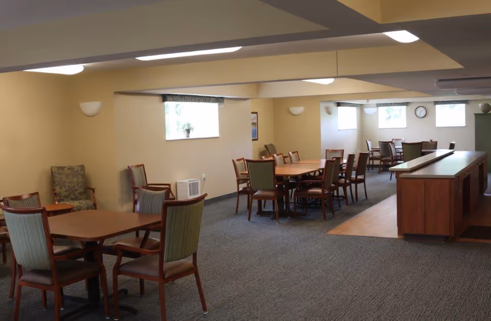 A spacious common area with multiple wooden tables and chairs arranged for group seating. The room has beige walls, carpeted flooring, several windows letting in natural light, and a wooden counter or bar area on the right side. There are also a few armchairs and small tables along the walls.