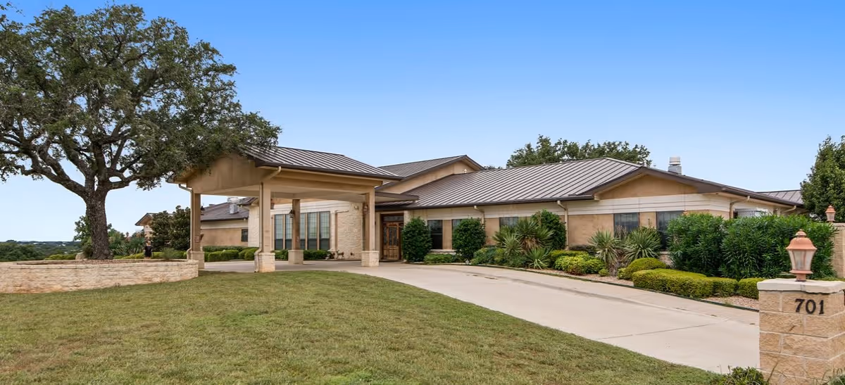 Exterior view of a single-story assisted living facility building with a covered entrance, surrounded by well-maintained landscaping including bushes, trees, and a lawn under a clear blue sky.