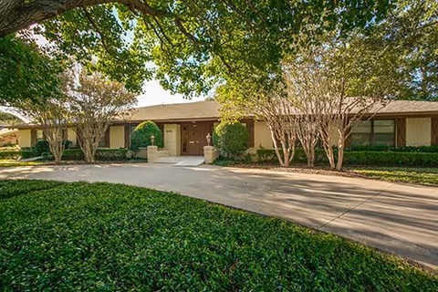 Single-story building with a flat roof surrounded by trees and shrubs, with a paved driveway and walkway leading to the entrance under a canopy of green leaves.