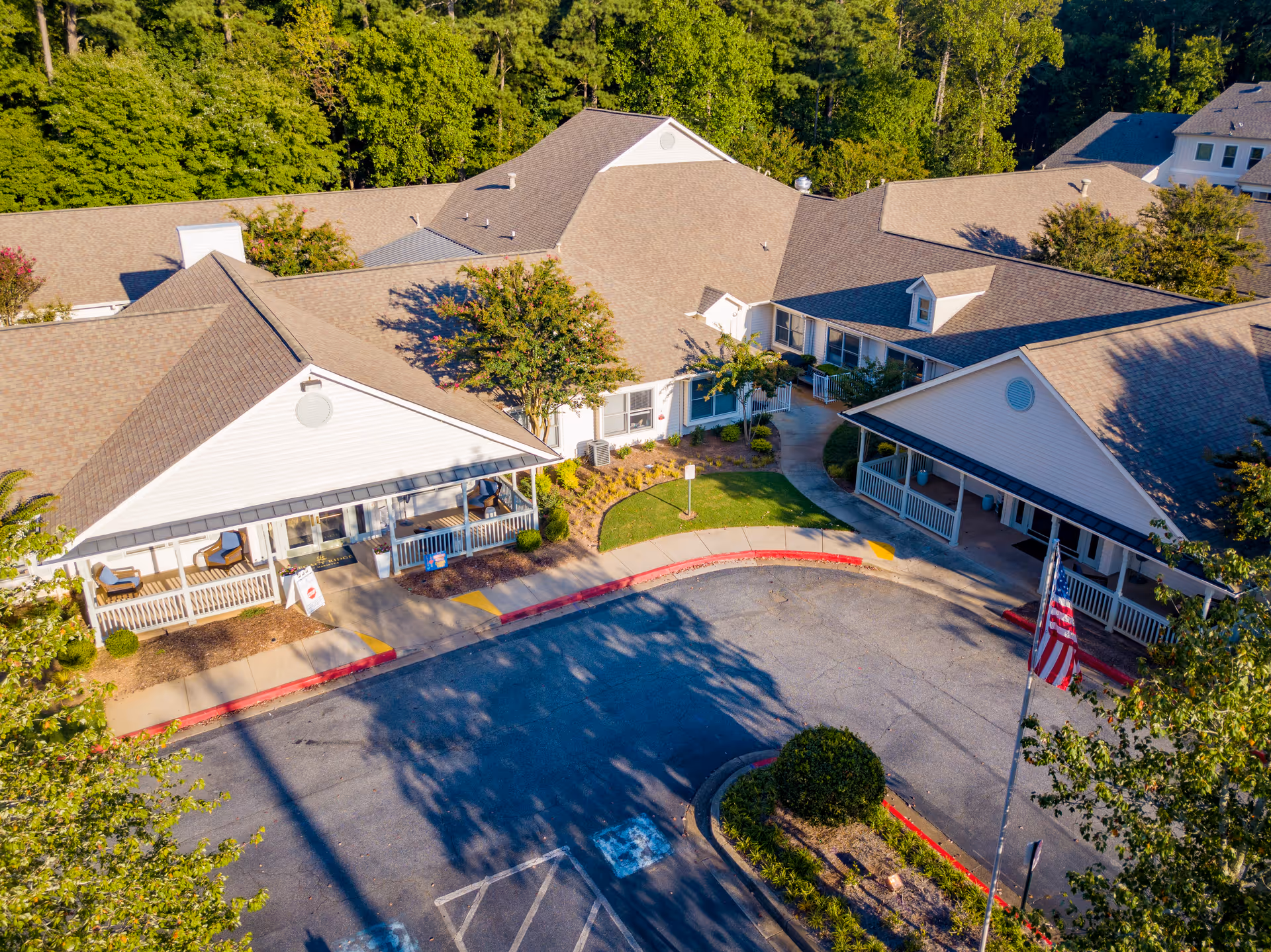 Aerial view of the front entrance of a senior living facility named Legacy Ridge at Marietta, showing a circular driveway with an American flag, landscaped greenery, and multiple connected buildings with beige roofs surrounded by trees.