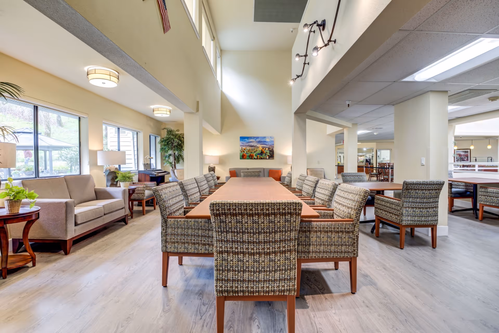A bright and spacious common area in a retirement community featuring a long rectangular table surrounded by patterned chairs. The room has large windows letting in natural light, a beige sofa with side tables and lamps, potted plants, and a colorful painting on the far wall. The flooring is light wood, and the ceiling has modern light fixtures.