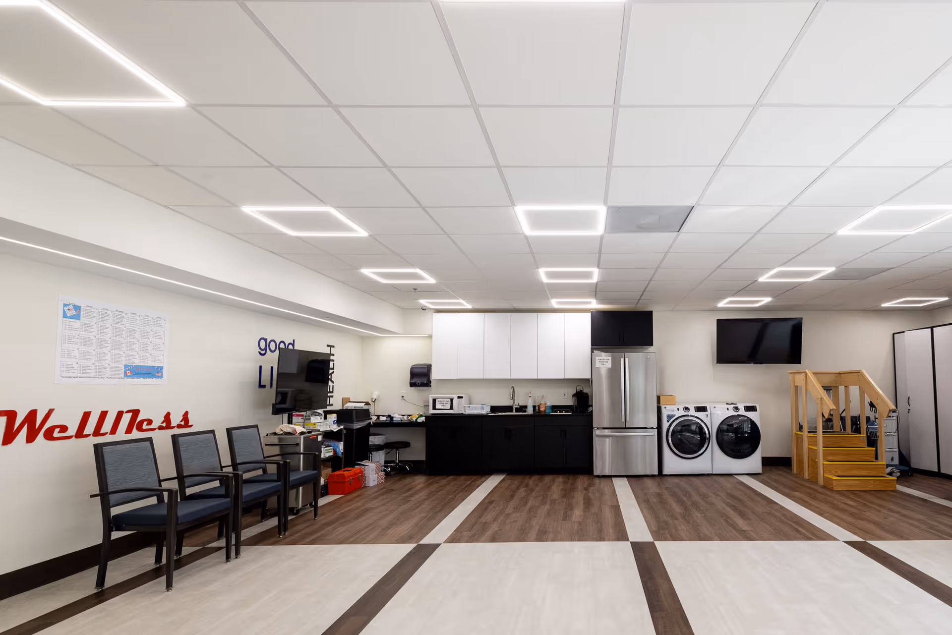 A spacious room with a kitchenette area featuring a refrigerator, microwave, sink, and cabinets. There are two front-loading washing machines next to a small wooden staircase with handrails. Several chairs are lined up against the wall on the left side, which has the word 'Wellness' displayed in red letters. The ceiling has modern square LED lights, and the floor has a patterned design with light and dark wood tones. A wall-mounted TV is visible above the washing machines.