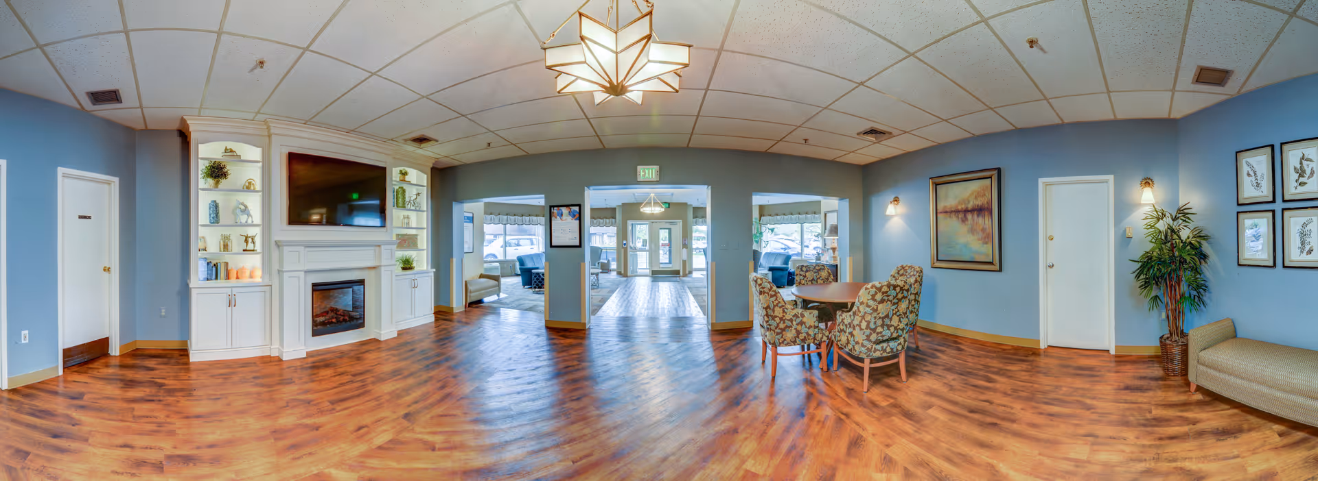 A spacious senior living facility common area with light blue walls and wood flooring. The room features a white fireplace with built-in shelves on either side, decorated with plants and ornaments. There is a flat-screen TV mounted above the fireplace. To the right, there is a small round table with four floral upholstered chairs, a framed painting on the wall, and a potted plant next to a beige bench. The ceiling has recessed lighting and a geometric light fixture. Double glass doors lead to another seating area with large windows allowing natural light to enter.