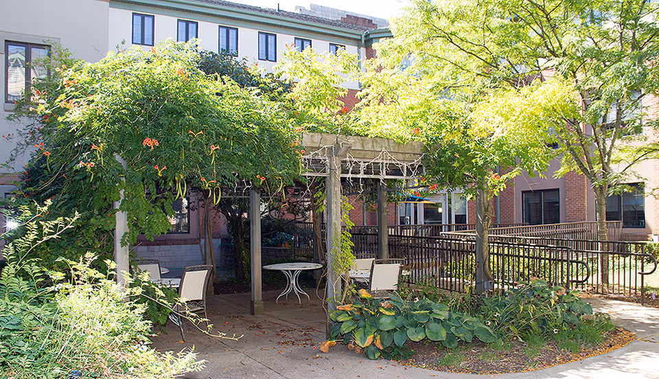 Outdoor seating area with a pergola covered in green vines and orange flowers, surrounded by trees and plants. Several chairs and a round table are placed under the pergola. The background shows a multi-story brick and beige building with windows.