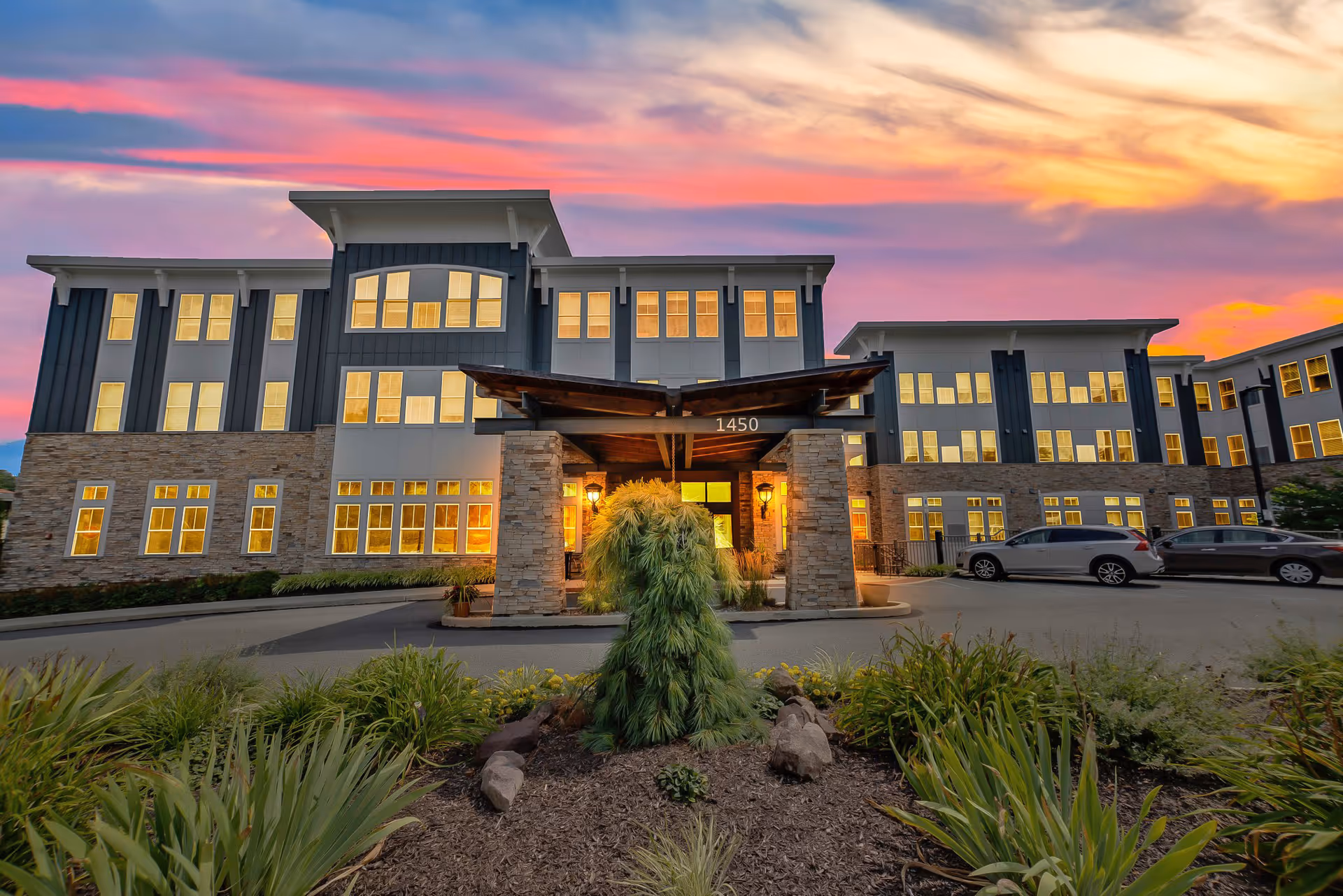 Front exterior view of a modern senior living facility building at sunset with warm lights glowing from the windows, a covered entrance with stone pillars, landscaped greenery, and parked cars.