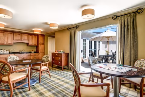 A bright and cozy common area with wooden cabinets and a kitchenette on the left, round tables with chairs featuring floral upholstery, and a sliding glass door leading to an outdoor patio with a table and umbrella.