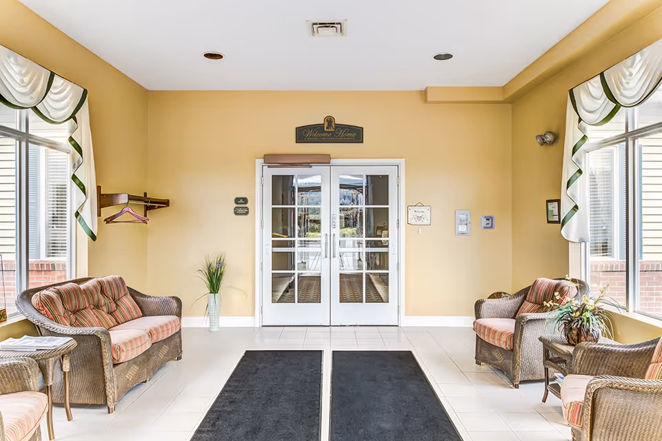 Entrance area of a senior living facility with double glass doors, two wicker armchairs with striped cushions, a wicker loveseat, a small wicker table with a plant, large windows with white and green valances, and a 'Welcome Home' sign above the doors.