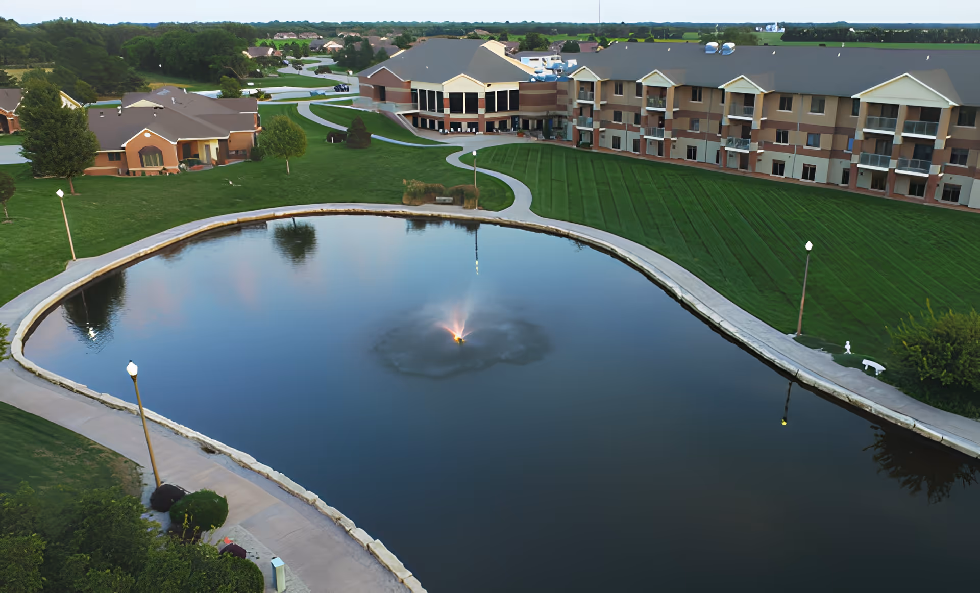 Aerial view of Schowalter Villa senior living facility showing a large pond with a central fountain, surrounded by a walking path and well-maintained green lawns. The multi-story residential buildings are visible in the background under a clear sky.