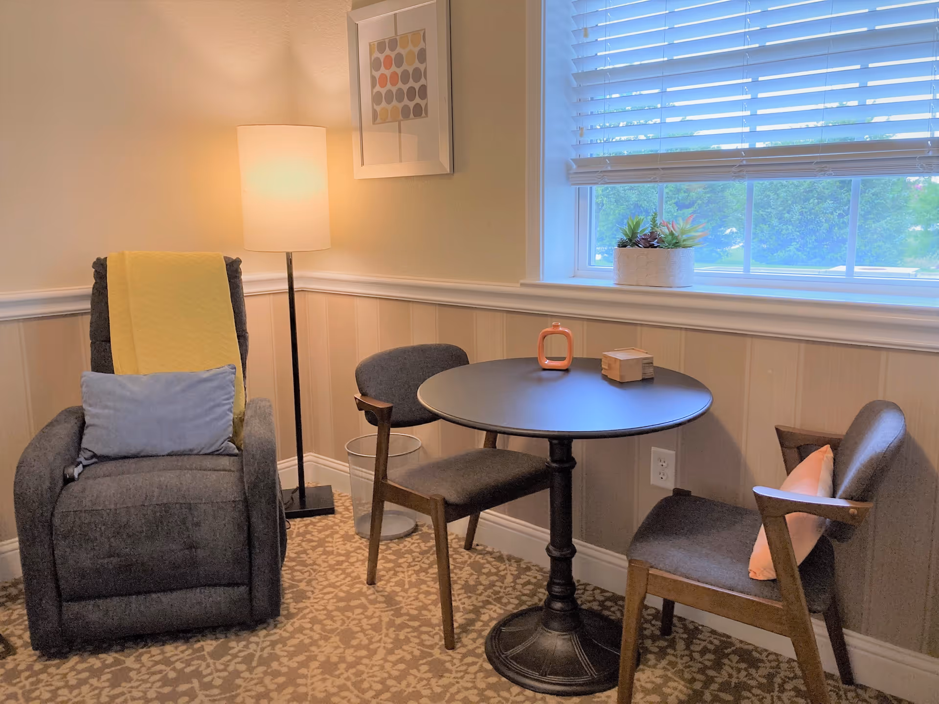 A cozy corner in a room with a dark gray recliner chair draped with a yellow blanket and a light blue pillow, a round black table with two gray cushioned chairs, a floor lamp with a white shade, a window with white blinds, and a small plant on the windowsill.