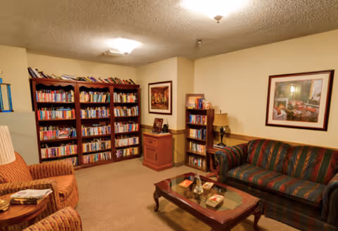 A cozy living room area with a striped sofa and two armchairs surrounding a wooden coffee table. The room features multiple bookshelves filled with books, framed artwork on the walls, and a table lamp on a small bookshelf. The walls are painted a light beige color and the carpet is brown.