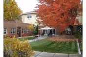 Outdoor courtyard area of a senior living facility with a large tree displaying red autumn leaves, green grass, a paved walkway, benches, and a building with large windows and a patio umbrella.