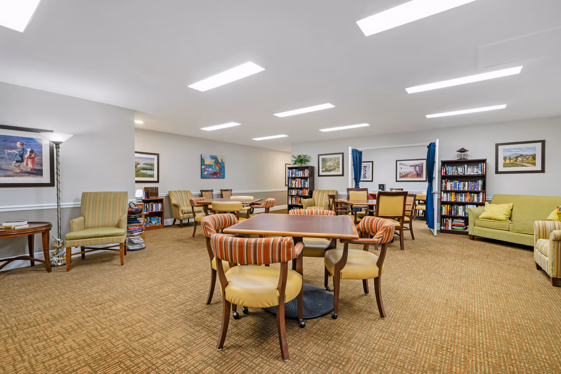 A bright communal lounge with tables, chairs, bookshelves, and sofas in a senior living facility.
