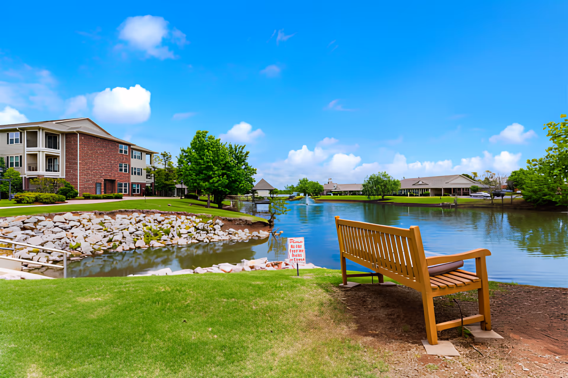 A wooden bench overlooks a pond with manicured lawns and a three-story brick-and-siding senior living building under a bright blue sky.