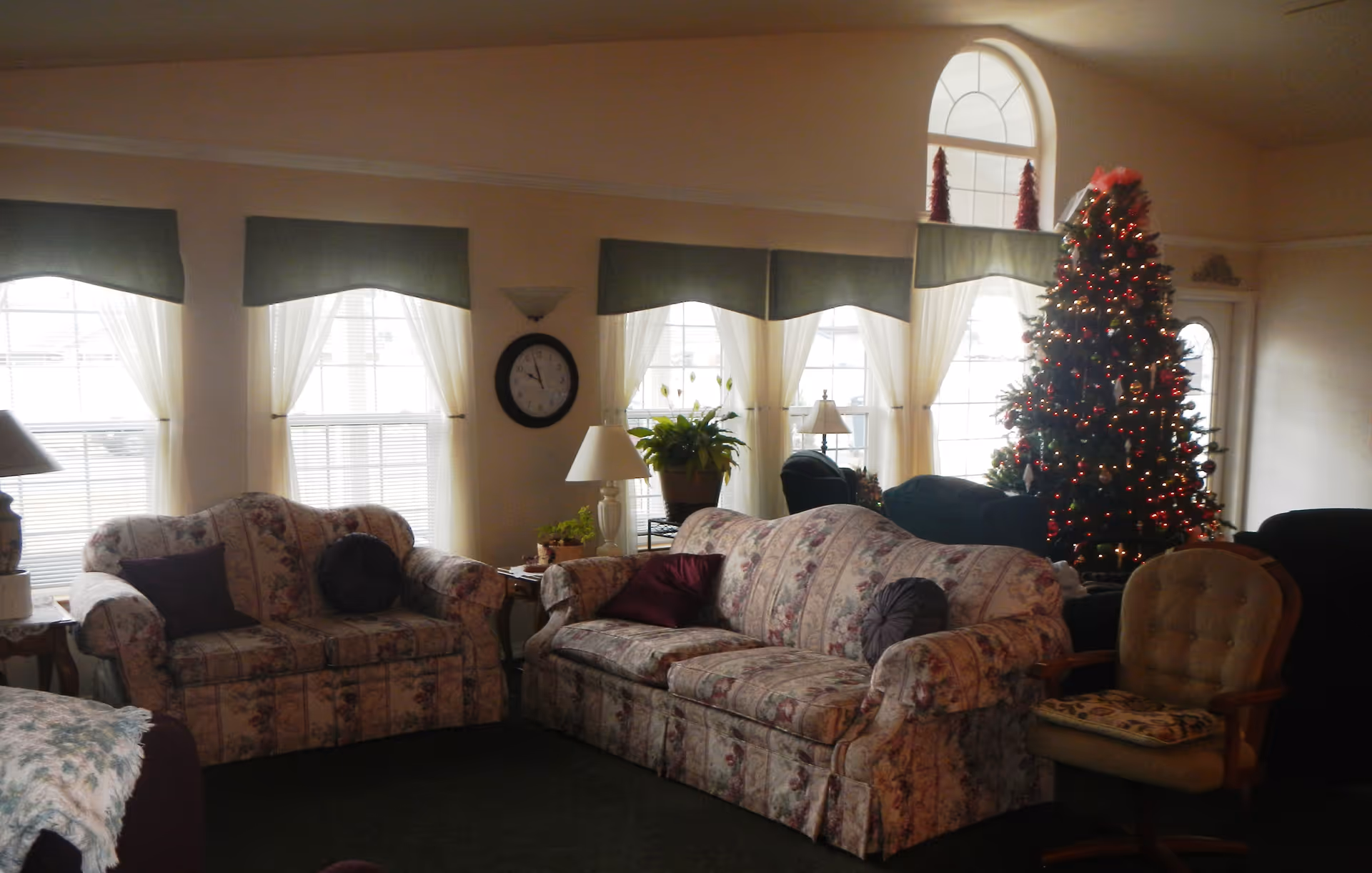 A cozy living room with floral patterned sofas, a decorated Christmas tree with red lights, several windows with sheer white curtains and green valances, a wall clock, table lamps, and a cushioned armchair.