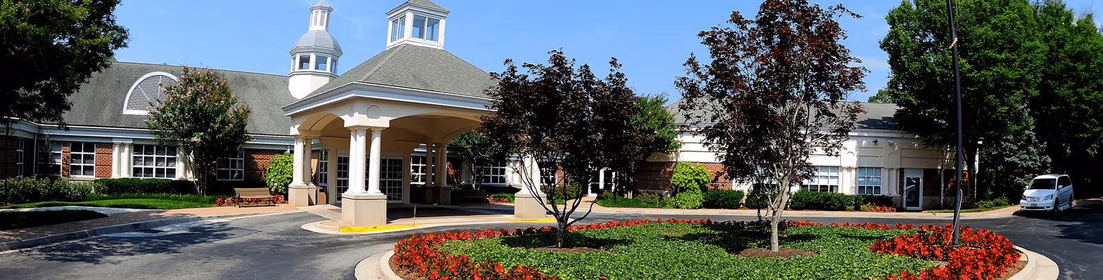 Front exterior view of Blakehurst facility showing a circular driveway with a landscaped center island featuring trees and red flowers, a covered entrance supported by white columns, and a brick building with multiple windows under a clear blue sky.