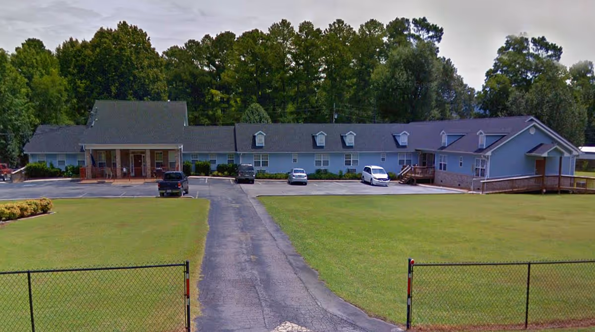 Single-story light-blue senior living building with a driveway, parking area and large front lawn set against trees.