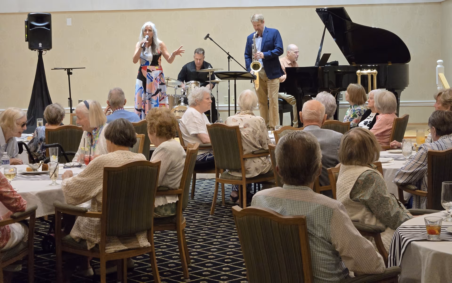 A small band performs on a stage while seniors sit at round dining tables in a community dining room.