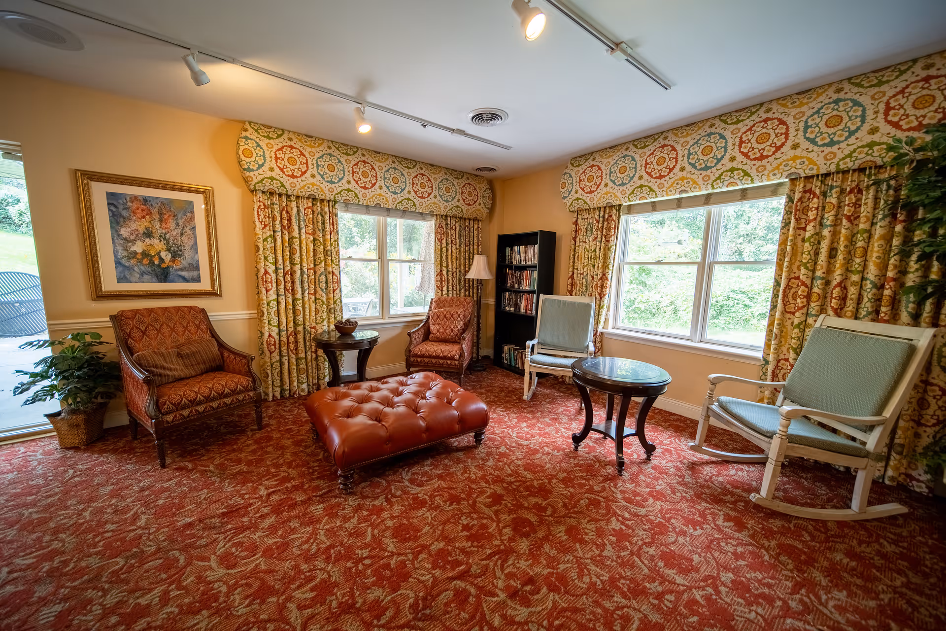 A cozy living room with patterned red carpet and matching curtains featuring a colorful circular design. The room has two large windows letting in natural light. There are two upholstered armchairs with a red and brown pattern, two light blue cushioned rocking chairs, a tufted red leather ottoman, and two small round wooden tables. A bookshelf filled with books and a floor lamp are placed near the windows. A framed floral painting hangs on the wall, and a potted plant is near the sliding glass door leading outside.