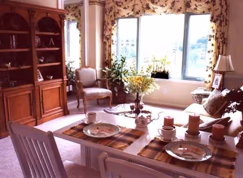 Sunlit dining room with a table set for meals beside large windows and plants.