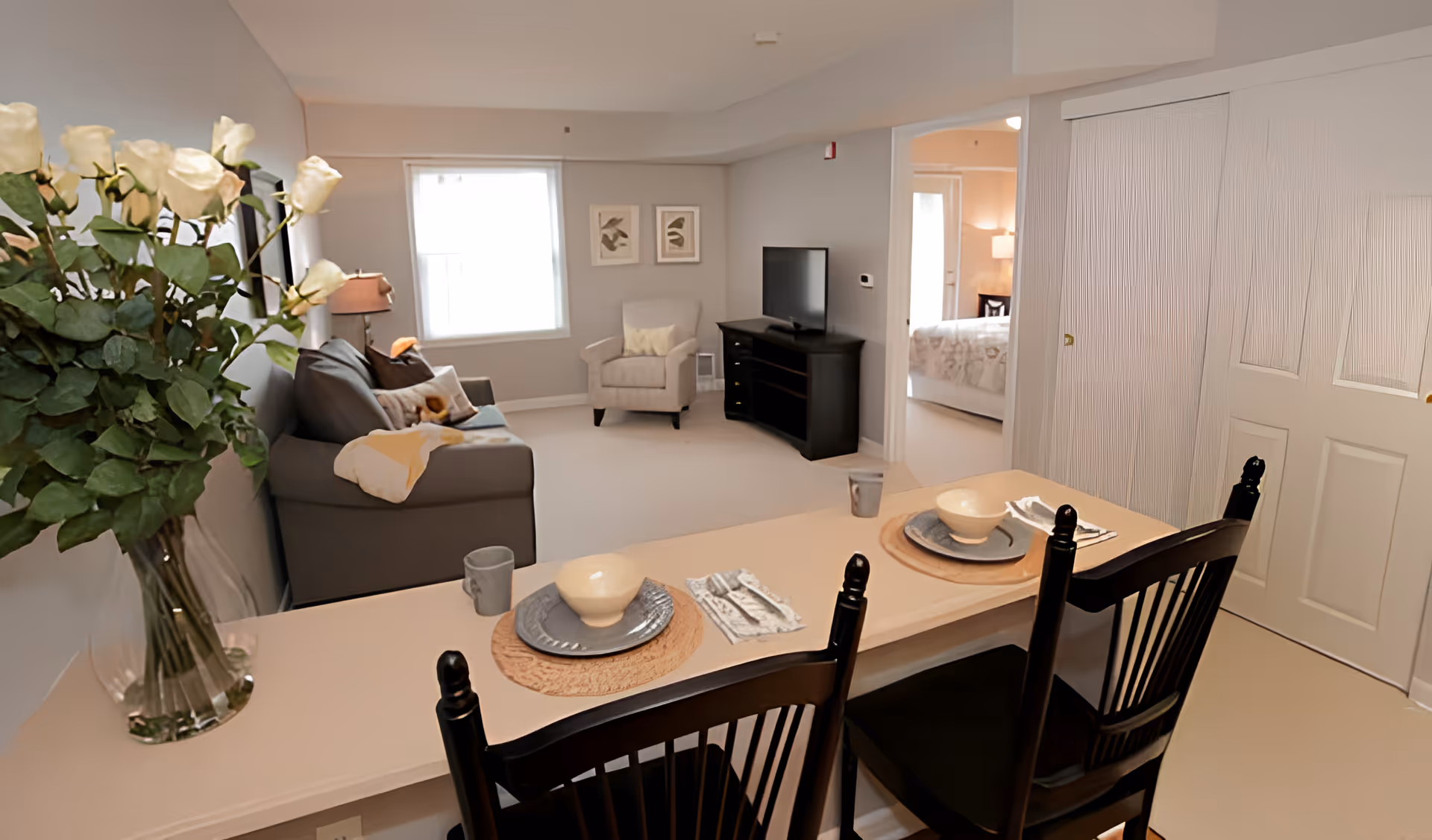 Interior view of a senior living facility showing a cozy living room with a gray sofa, an armchair, a TV on a black stand, and a window with white curtains. In the foreground, there is a counter with two place settings including bowls, plates, napkins, and mugs, along with two black chairs. A vase with white roses is placed on the counter. A bedroom is visible through an open doorway.