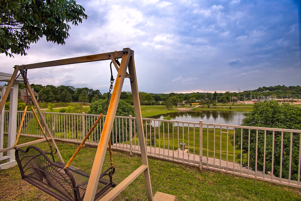 A metal porch swing on a grassy balcony overlooking a fenced lawn, pond, and trees under a cloudy sky.
