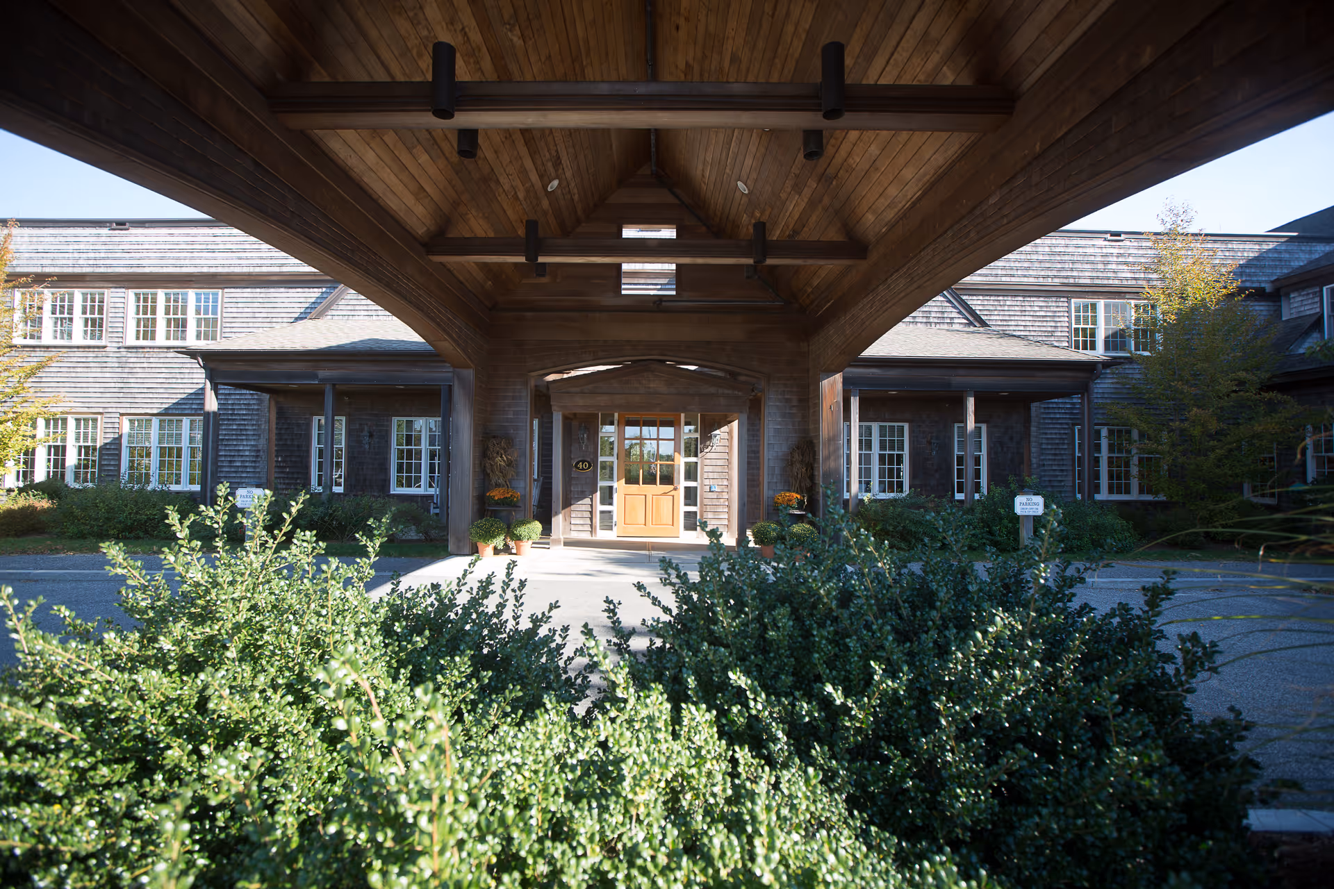 Entrance of Sherburne Commons facility with a wooden covered driveway, surrounded by green bushes and trees, and a building with multiple windows in the background.