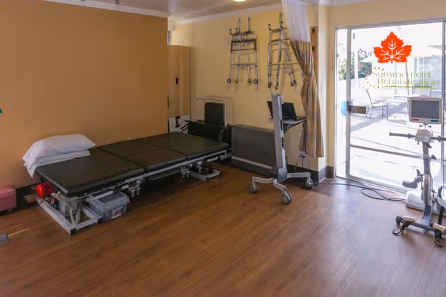 A rehabilitation room with a padded treatment table, walkers hanging on the wall, a computer on a mobile stand, and exercise equipment near a glass door leading to an outdoor patio area.