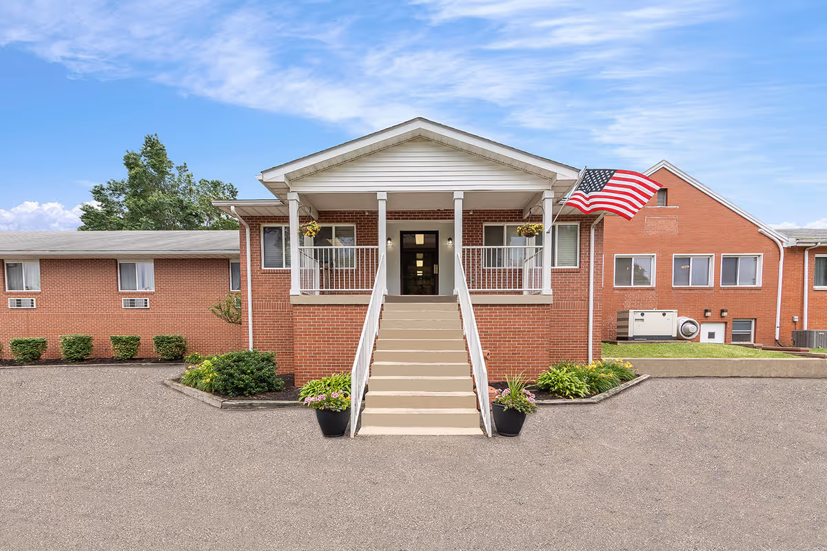 Front entrance of a red brick senior living facility with stairs leading to a covered porch, an American flag, and surrounding landscaping.