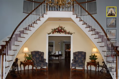 Interior view of a senior living facility featuring a grand double staircase with wooden steps and white railings curving upwards on both sides. Below the staircase, there are two upholstered armchairs with floral patterns, each accompanied by a wooden side table with a lamp and a potted plant. A floral arrangement decorates the wall above the doorway in the center, leading to another room with visible furniture.
