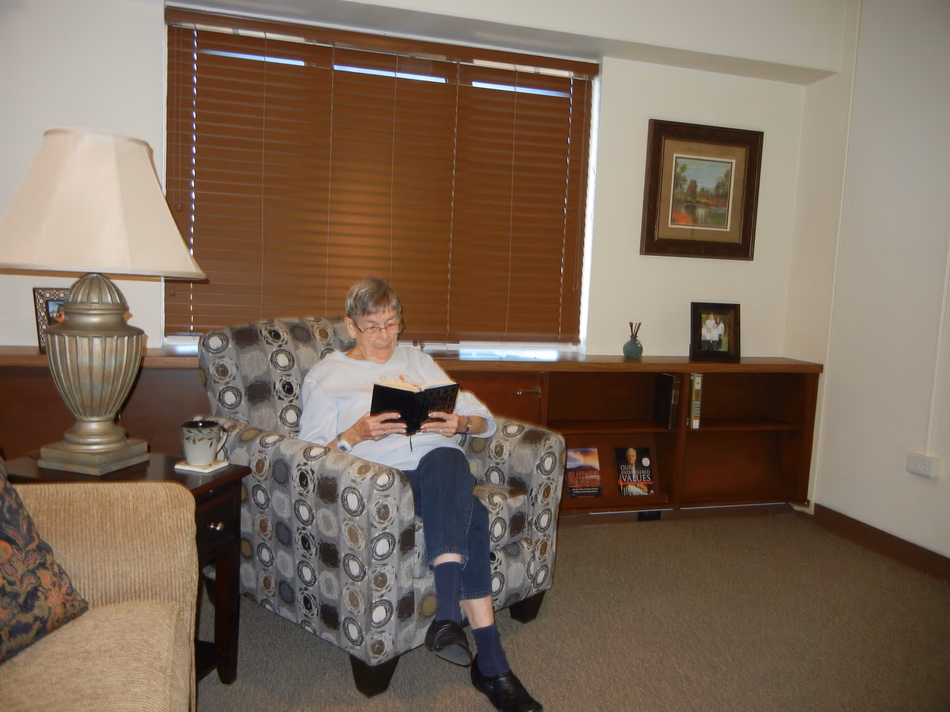 An elderly woman sitting in a patterned armchair reading a book in a cozy living room. The room has brown blinds covering the window, a wooden side table with a lamp and a cup, a beige sofa with a patterned pillow, and a wooden bookshelf with a few books and framed pictures on top.