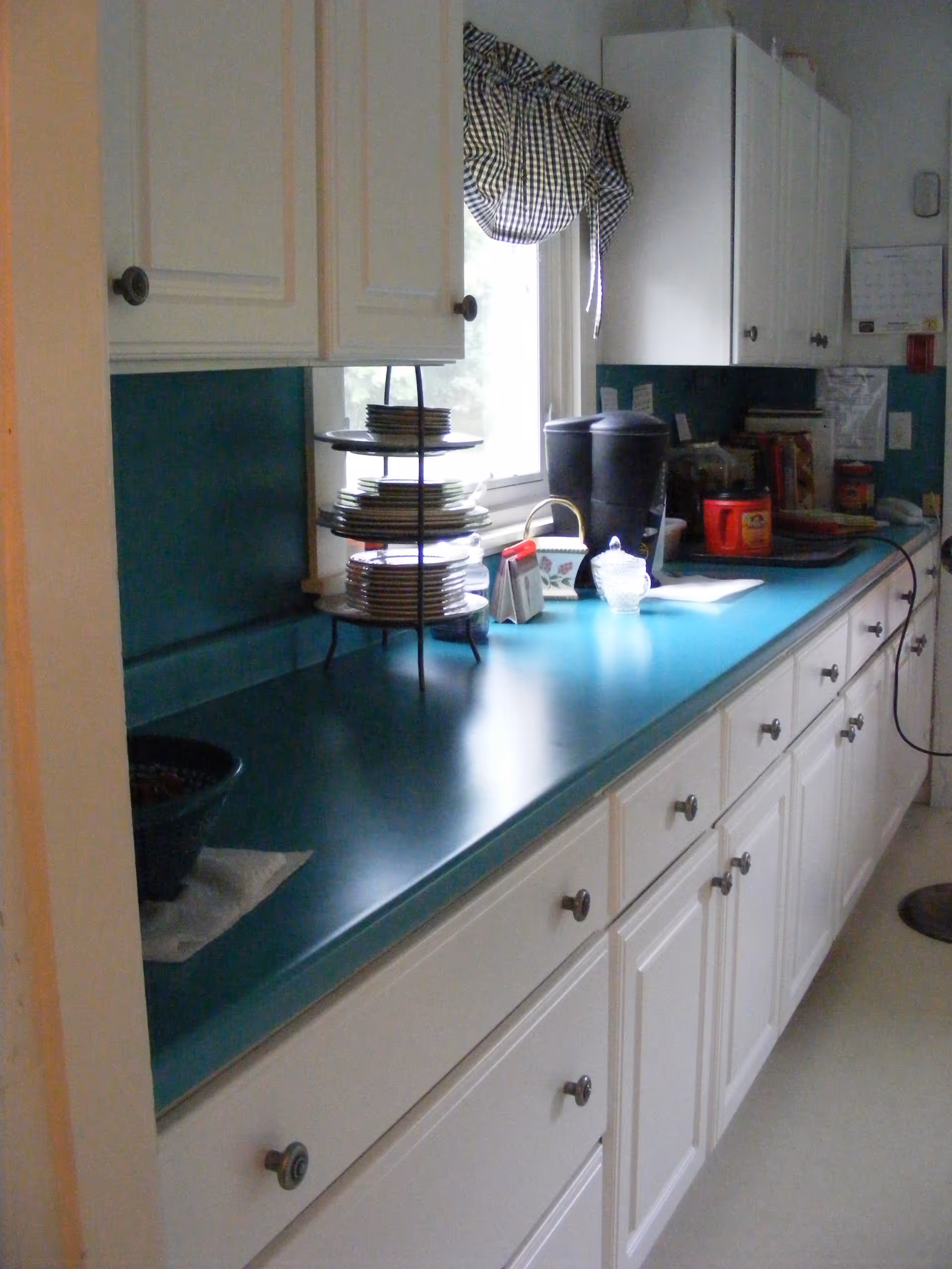 A kitchen counter with teal countertop and white cabinets above and below. On the counter, there is a tiered stand holding plates, a coffee maker, a sugar bowl, and various other kitchen items. A window with a checkered curtain is above the counter.