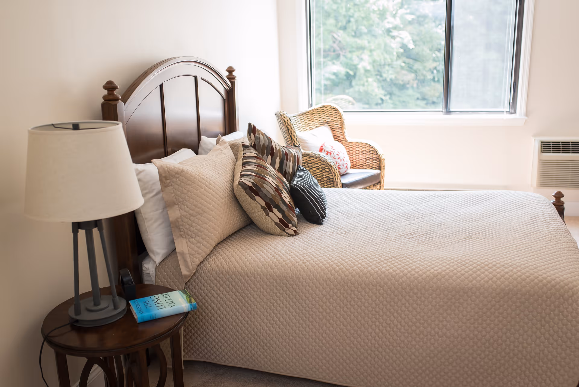 A neatly made bed with a wooden headboard, beige quilted bedspread, and several decorative pillows. Next to the bed is a round wooden side table with a lamp and a book titled 'The Long Letter'. In the background, there is a wicker chair with cushions near a large window showing greenery outside.