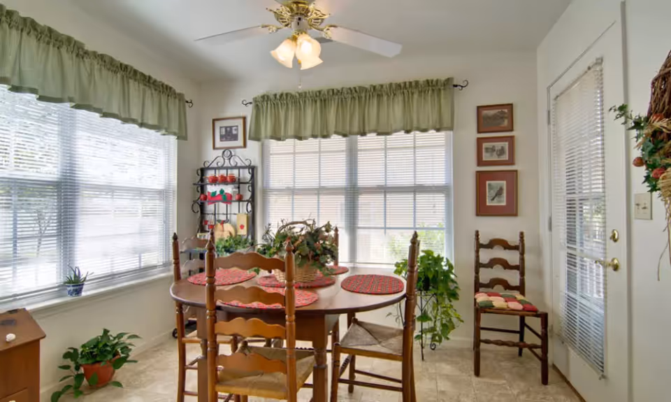 A bright dining area with a round wooden table surrounded by four wooden chairs with woven seats. The table is decorated with red placemats and a centerpiece of greenery. Large windows with green valance curtains let in natural light. There is a black metal shelf with decorative items, potted plants on the floor, framed pictures on the wall, and a door with blinds on the right side.