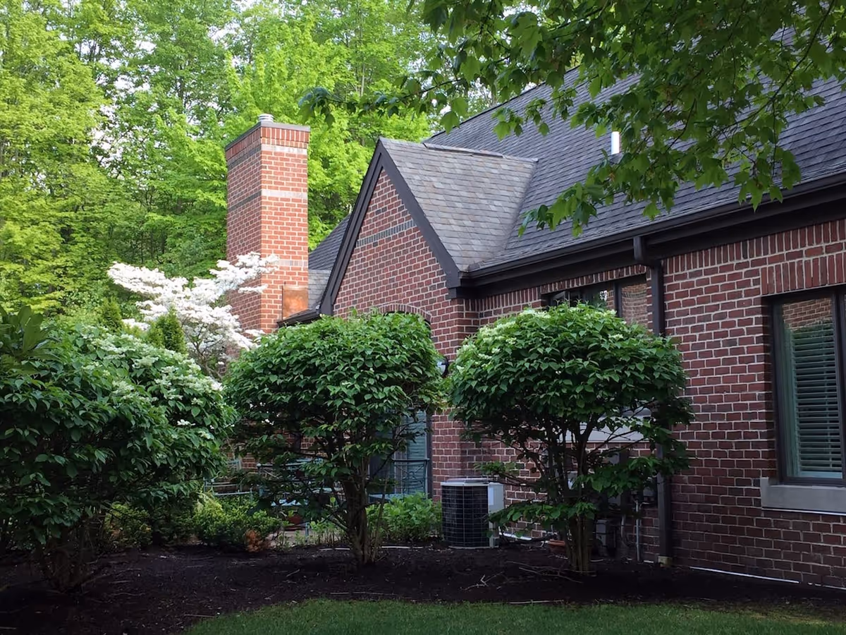 Brick building exterior with a shingled roof and chimney, surrounded by trimmed shrubs and green trees.
