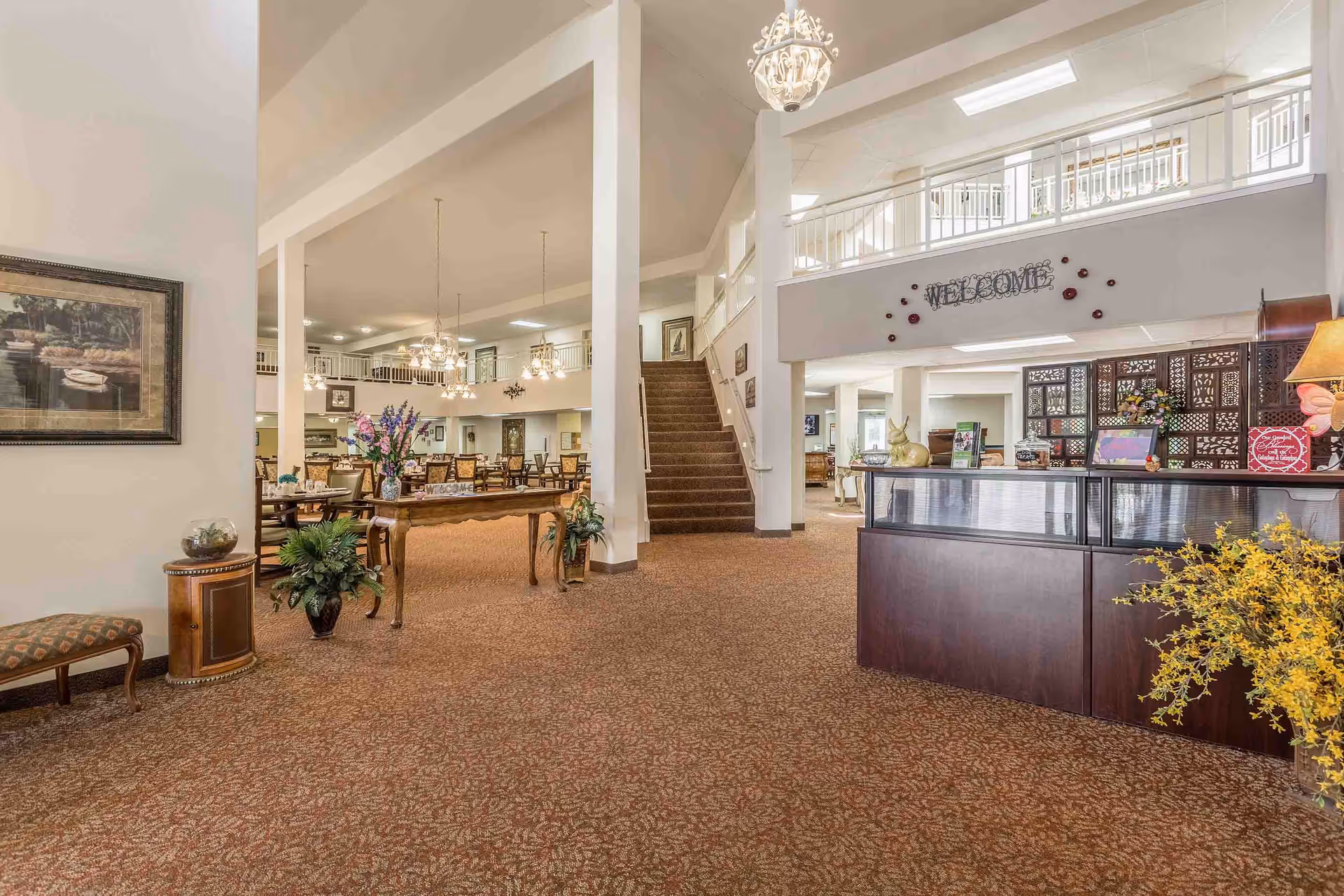 Spacious senior living facility lobby with a reception desk, central table of flowers, seating area and carpeted stairs to an upper balcony.
