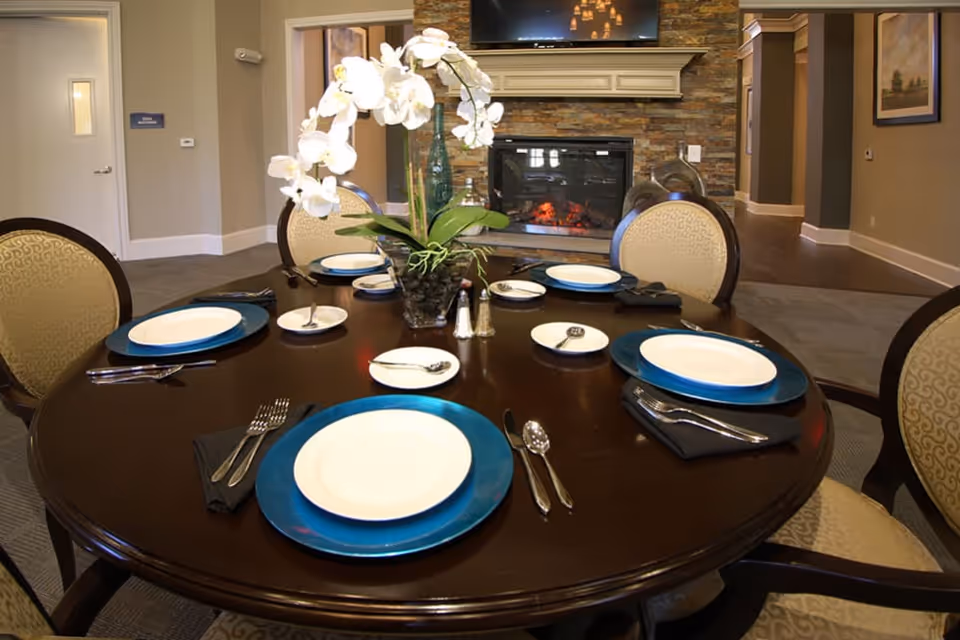A round dining table set for six with white plates on blue chargers, silverware, and black napkins. A vase with white orchids is in the center of the table. In the background, there is a stone fireplace with a TV mounted above it and beige walls with framed artwork.