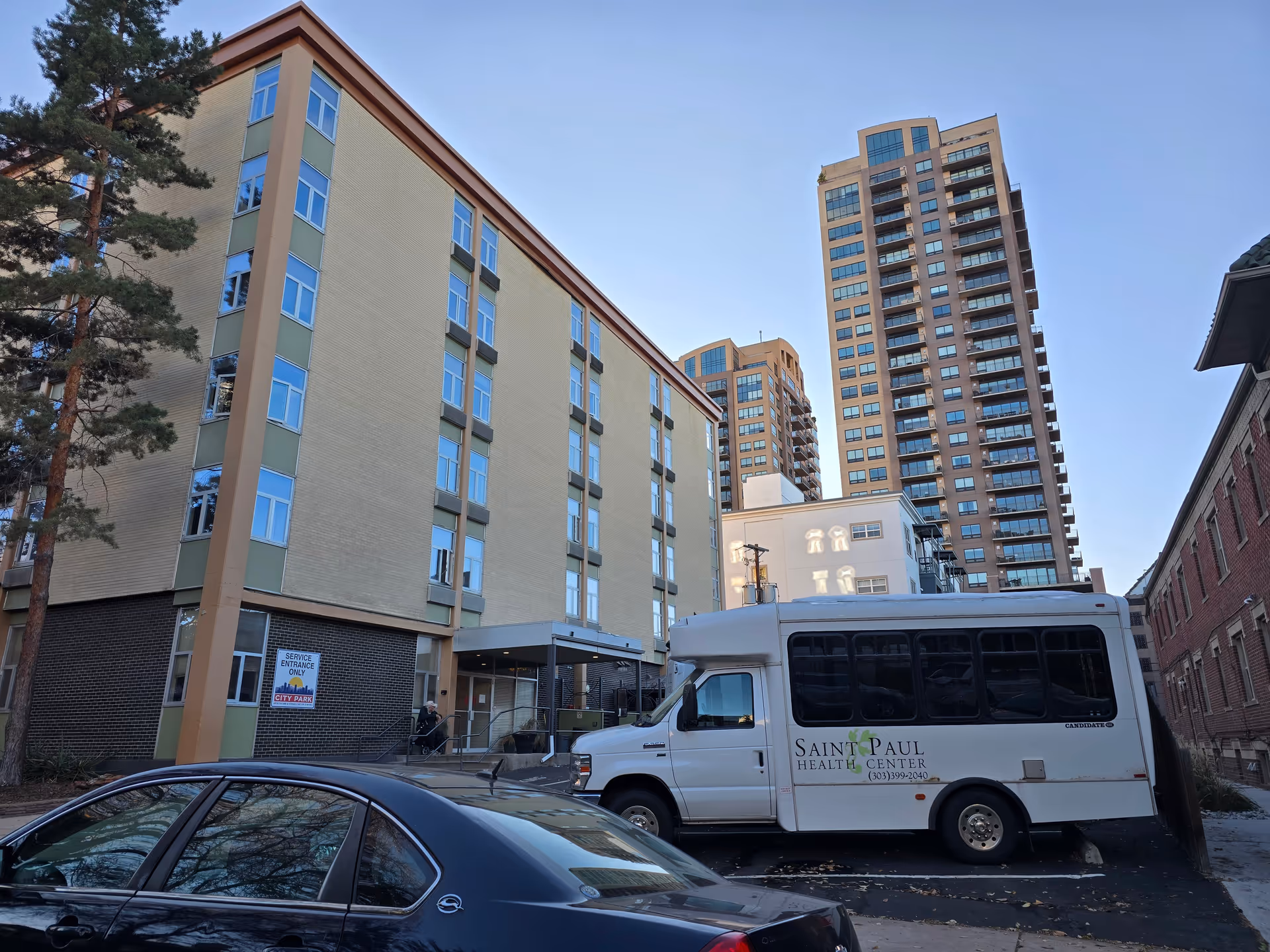 Exterior view of St Paul Health Center, a multi-story building with a beige and green facade. A white shuttle bus with the St Paul Health Center logo and phone number is parked in front. There are other tall buildings in the background and a black car parked in the foreground.