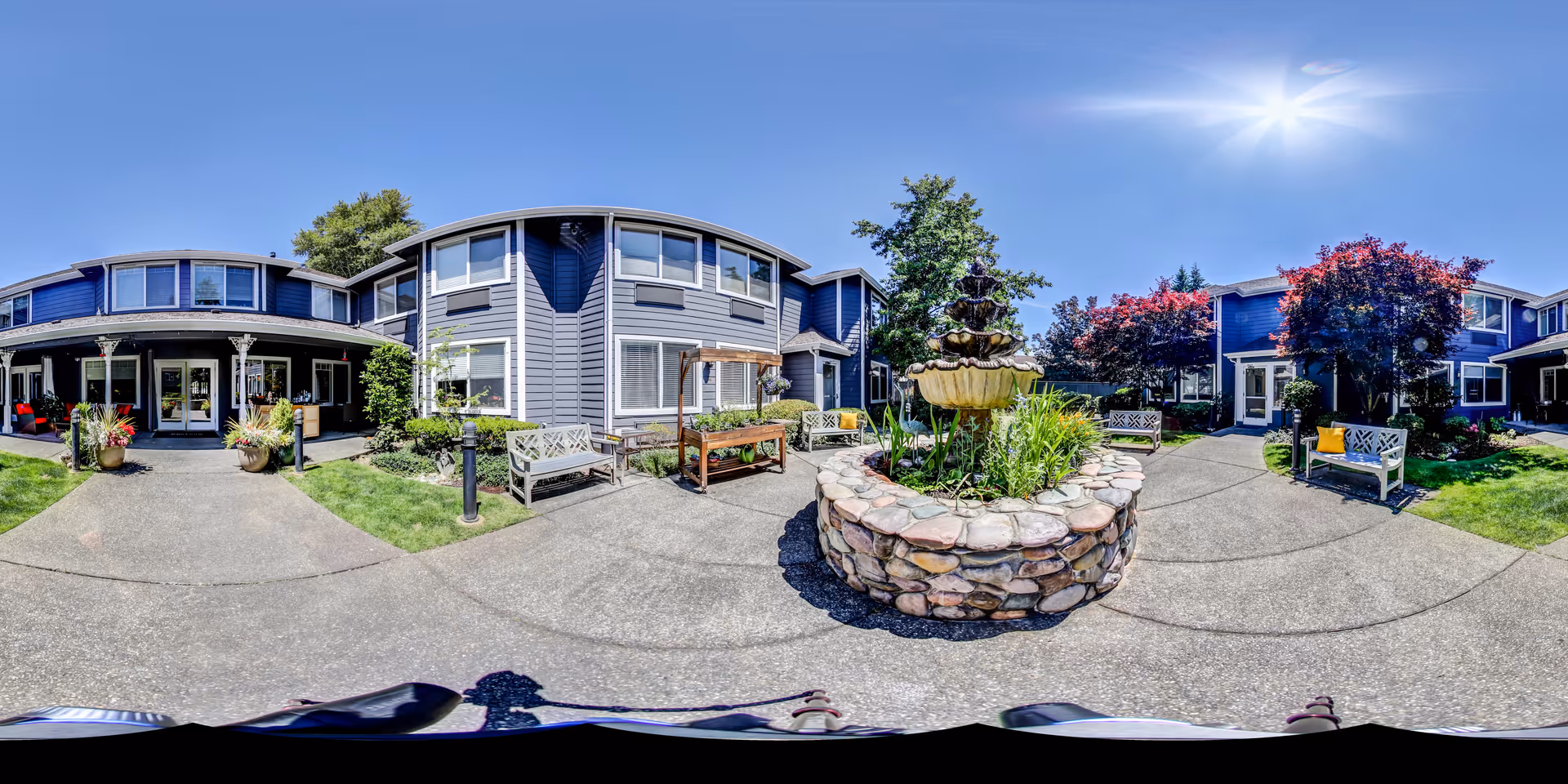 Outdoor courtyard area of a senior living facility with a stone fountain in the center surrounded by benches, plants, and trees. The building is two-story with blue siding and white trim under a clear blue sky with the sun shining brightly.